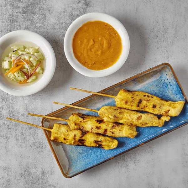 Grilled chicken satay skewers on a blue plate with peanut sauce and cucumber salad.